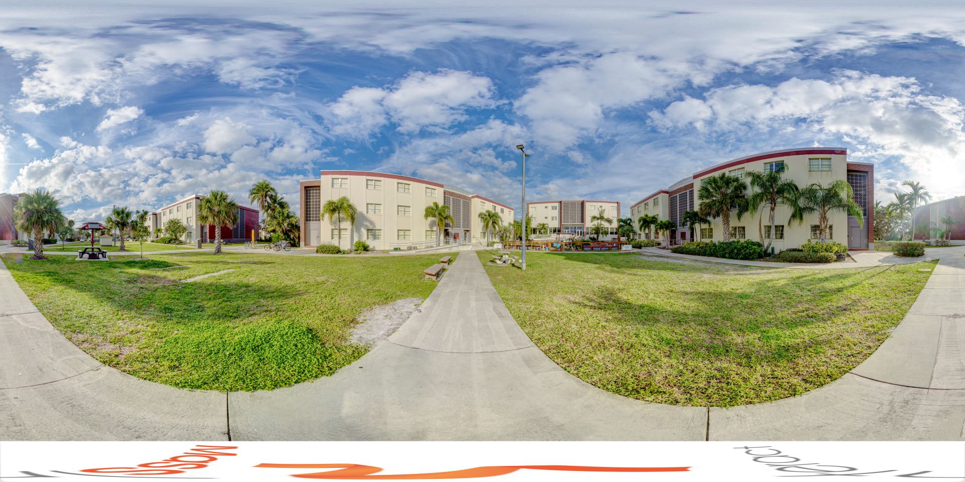 Panoramic view of a residential college campus with modern buildings, palm trees, and walkways under a vivid blue sky with clouds.